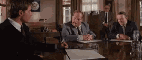 a group of men in suits and ties are sitting around a table talking to each other .
